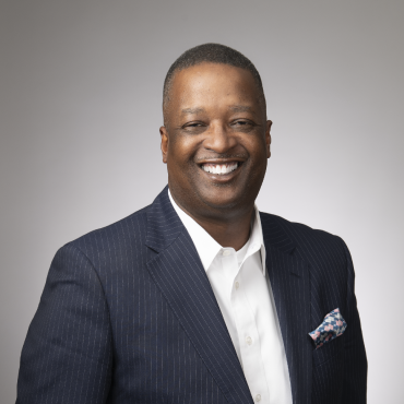 A professional portrait of a smiling man in a navy pinstripe suit and white shirt, standing against a gray background.